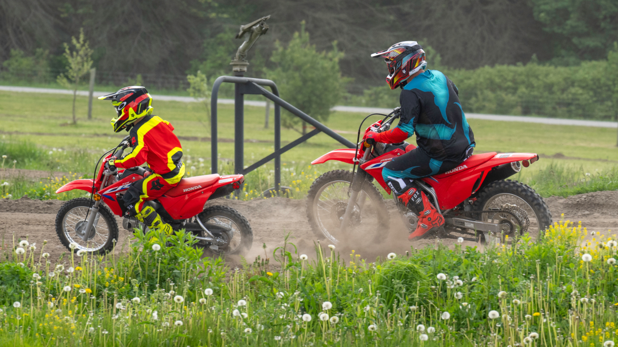 Young rider in front of adult rider on dirt track. Grass in foreground. Fence and field in background