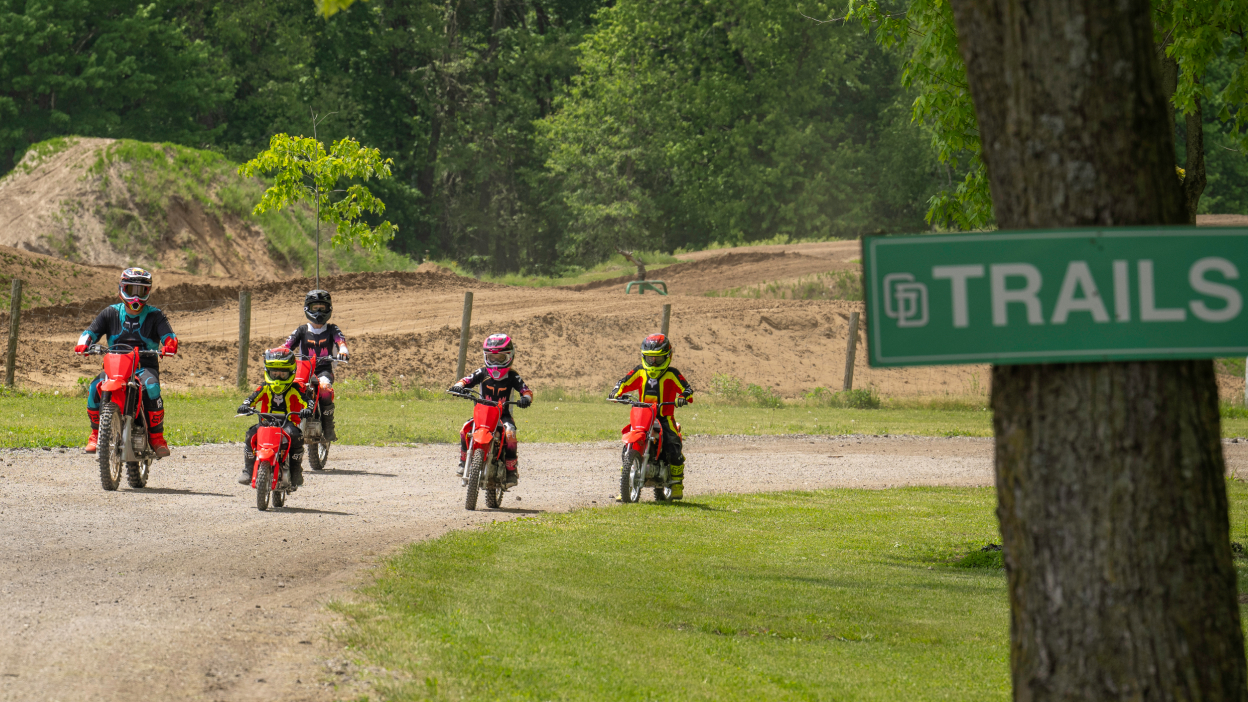 Two adult riders and three young riders front facing on track. Green Trails sign on tree to the right.