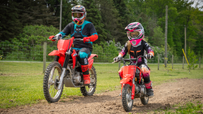 Adult rider and  young rider going right on dirt track. Green field, trees and fence in background.