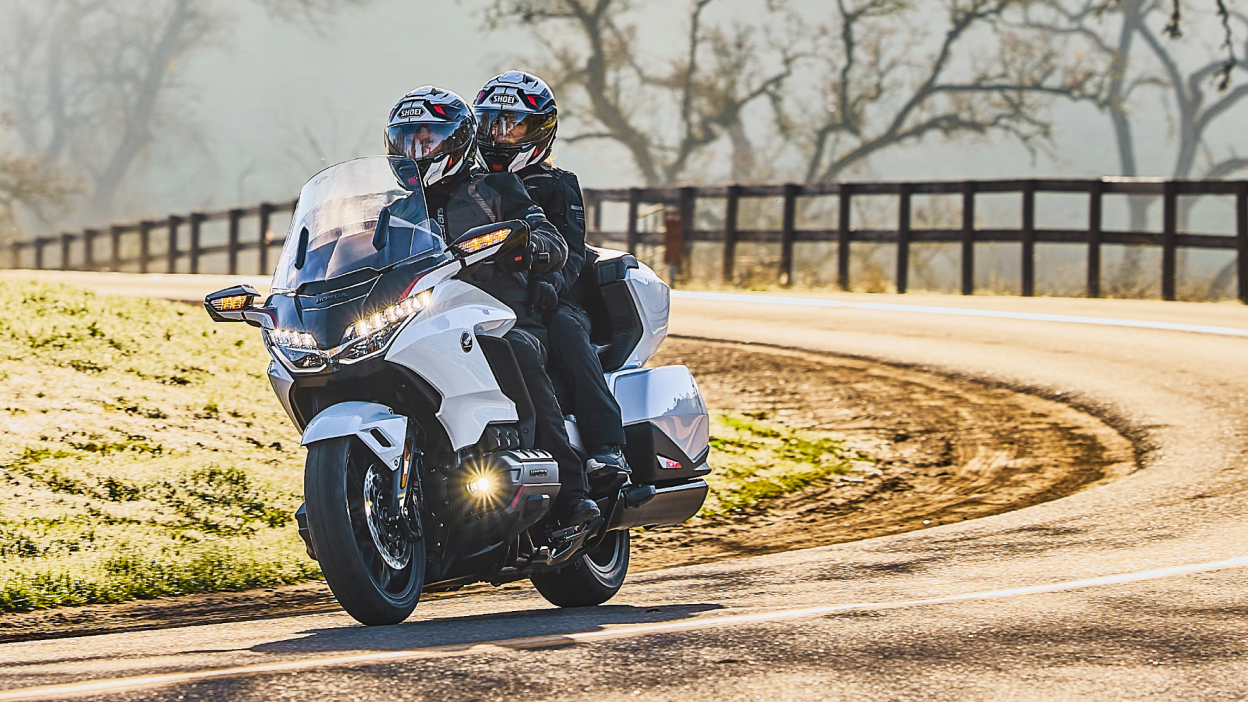 Two riders on white bike going left. Trees and fence in background.