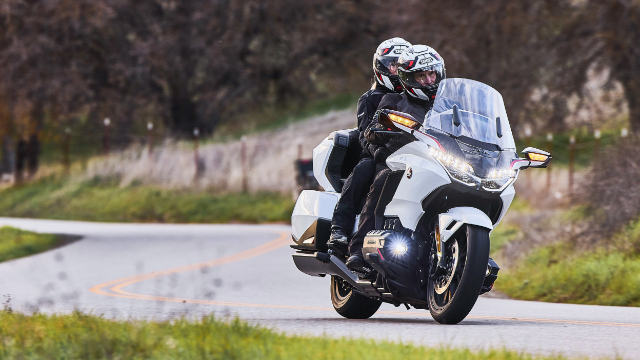 Two riders on white bike going right. Trees in background.