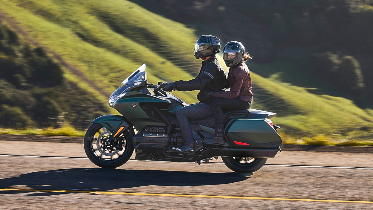Two riders on green bike going left. Passenger looking left. Green hill in background.