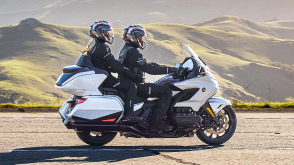 Two riders on white bike going right. Green Hills in background.