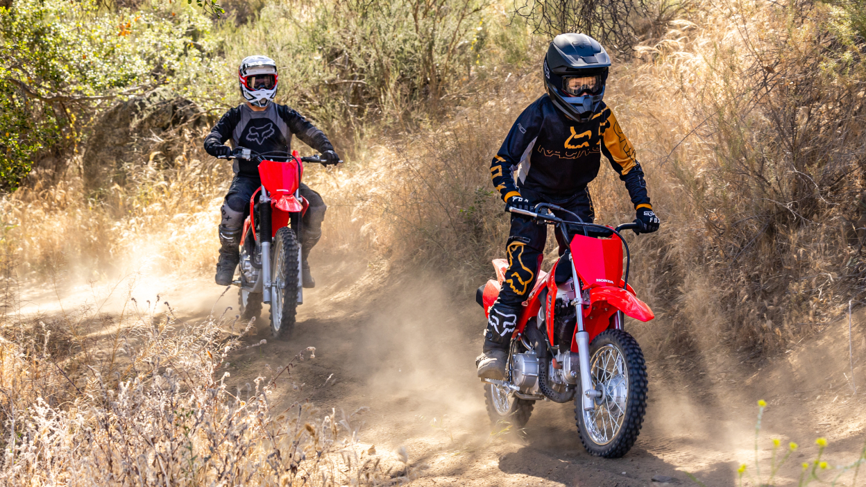 One young rider sitting up on bike on dirt track. One adult rider behind.