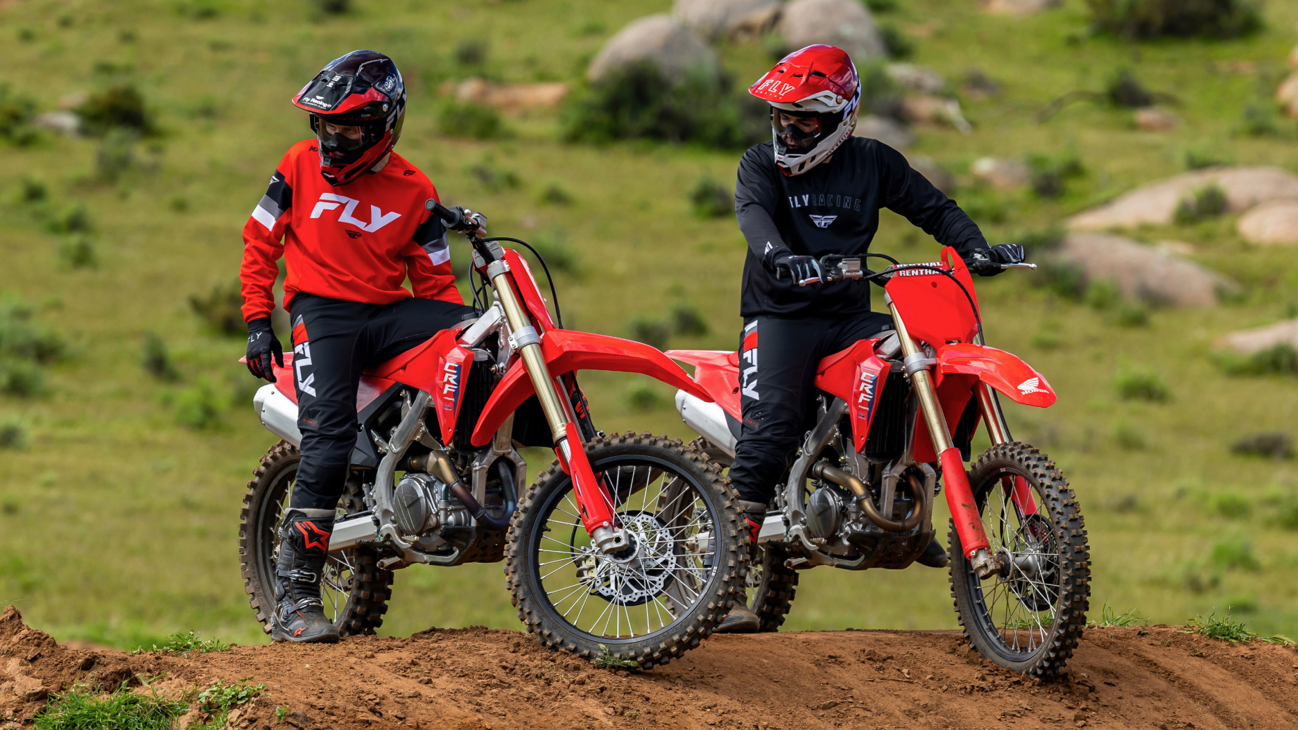 Two riders sitting on bikes on dirt hill. Green grass and grey rocks in background.