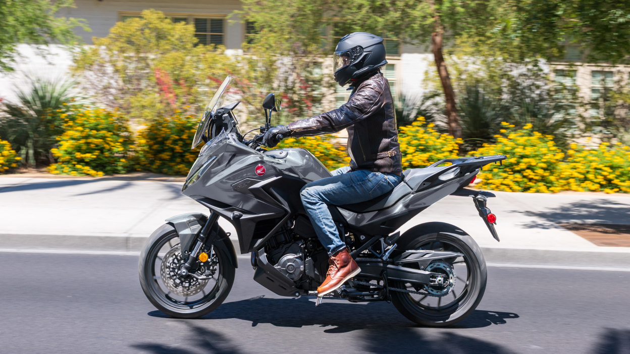 Rider on NT1100 going left on road. Pavement,  colourful bushes and small trees in background.