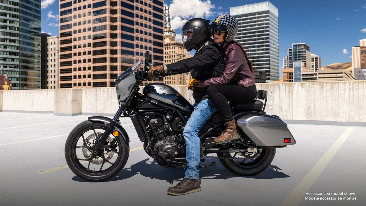 Two riders going right on street. Building and trees in background. Accessorized model shown.