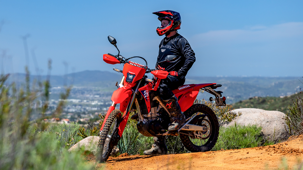 Pilote à l'arrêt sur une CRF450L. Paysage urbain et ciel bleu en arrière-plan.