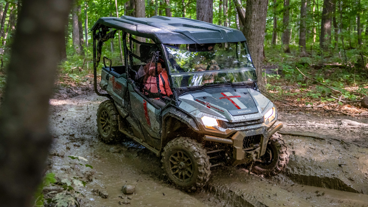 Pioneer going right through wet, muddy road. Forest in background. Headlights are on.